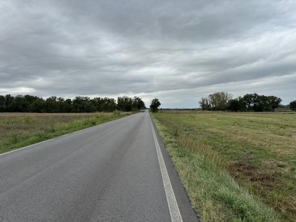 Tratto ciclovia su strada asfaltata in un’area rurale, affiancata da campi aperti. Alberi densi a sinistra, paesaggio aperto a destra